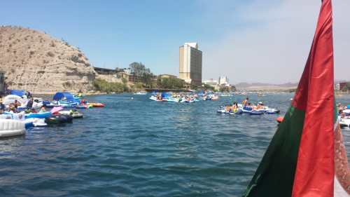 A busy river scene with people on various inflatable boats, surrounded by hills and a tall building in the background.
