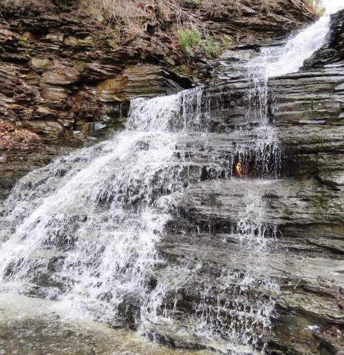 A cascading waterfall flows over rocky steps, surrounded by natural greenery and textured stone.