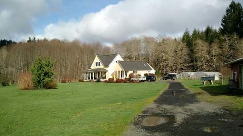 A spacious yellow house with a porch, surrounded by green grass and trees, under a partly cloudy sky.