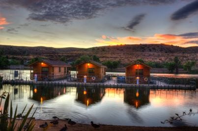 Sunset over a calm lake with wooden cabins reflecting on the water, surrounded by hills and ducks in the foreground.