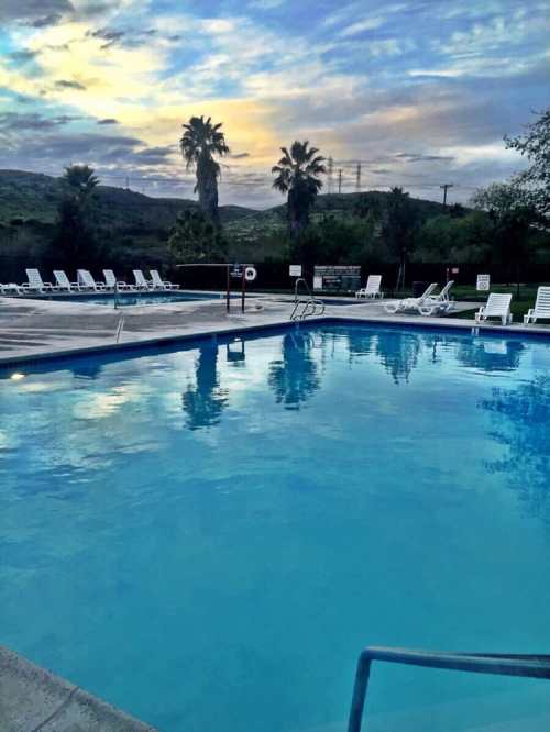 A serene swimming pool surrounded by lounge chairs, with palm trees and a colorful sunset in the background.