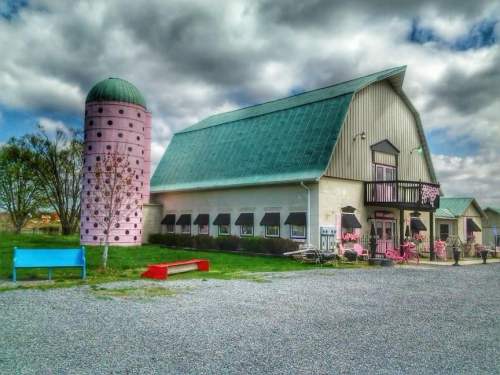 A colorful barn with a green roof and pink silo, surrounded by grass and a gravel path under a cloudy sky.