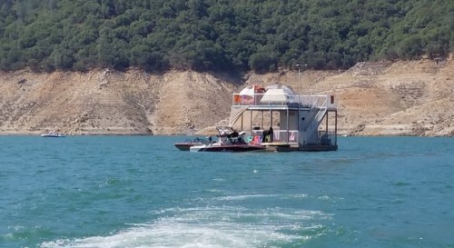 A floating house on a lake, surrounded by green hills and rocky shores, with a boat nearby.
