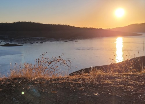 Sunset over a calm lake, with boats in the distance and silhouettes of hills against a golden sky.