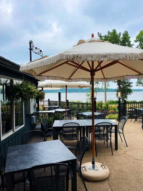 Outdoor dining area with tables under a large umbrella, overlooking a lake and surrounded by greenery.