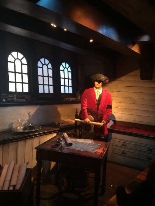 A pirate figure stands in a dimly lit cabin, with a table, map, and windows in the background.