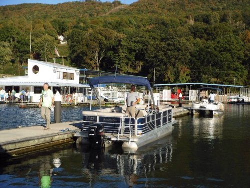 A pontoon boat is docked at a marina with people walking along the pier and a scenic hillside in the background.