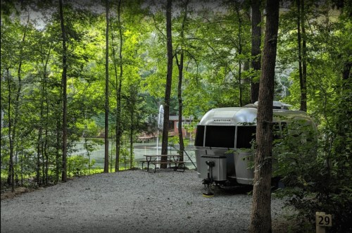 A silver Airstream trailer parked in a wooded area, surrounded by trees and gravel, near a serene lake.