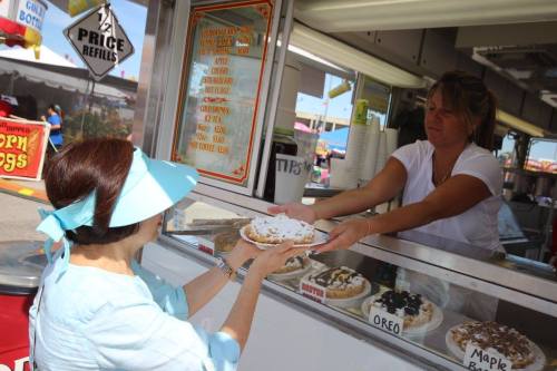 A woman in a sun hat receives a dessert from a vendor at a food stand, with various treats displayed.