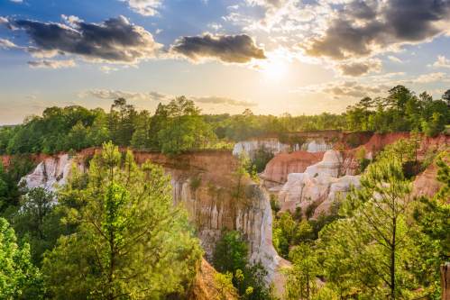 Sunset over a scenic canyon with layered cliffs and lush green trees under a partly cloudy sky.