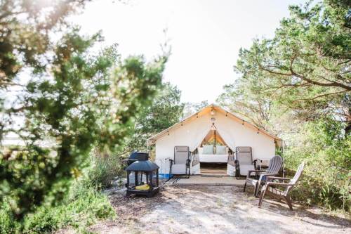 A cozy glamping tent surrounded by greenery, with chairs and a fire pit in front, inviting relaxation in nature.