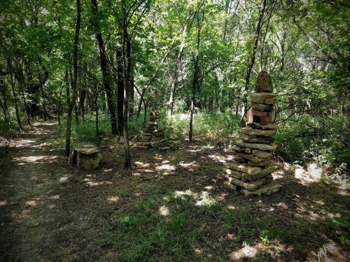 Two stone cairns stand among trees in a lush, green forest, with a dirt path visible on the left.