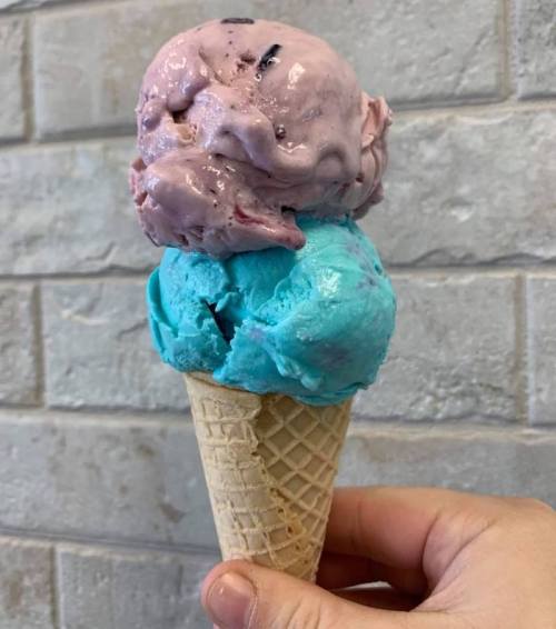 A hand holding a cone with two scoops of ice cream: one pink and one blue, against a brick background.
