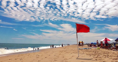 A beach scene with a red flag, people enjoying the sun, and fluffy clouds in a blue sky. Waves gently lap the shore.