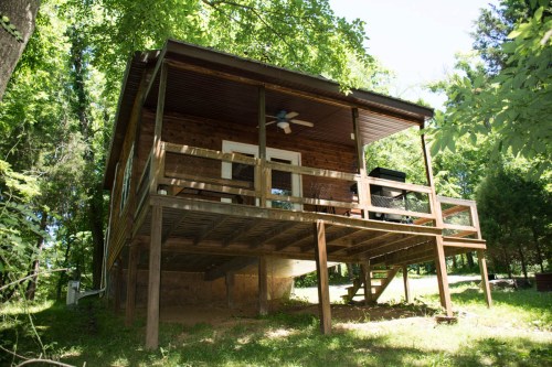 A wooden cabin on stilts surrounded by greenery, featuring a porch and a ceiling fan.