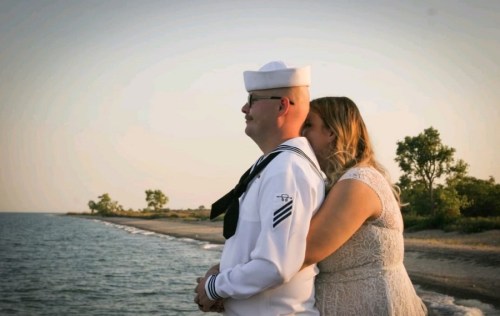 A couple stands by the water, with the woman embracing the man in a naval uniform, both gazing at the horizon.