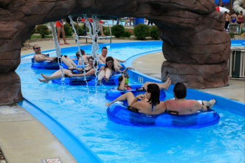 A group of people relaxes on blue inner tubes in a lazy river at a water park, surrounded by rocks and water features.