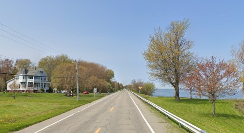 A scenic road lined with trees and a house, leading towards a lake under a clear blue sky.