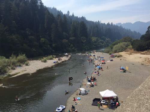 A sandy riverbank with people swimming, lounging, and enjoying a sunny day surrounded by trees and mountains.