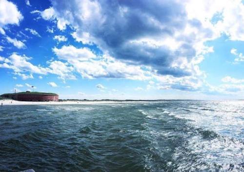 A scenic view of a beach with gentle waves, under a bright blue sky with fluffy clouds and a historic building in the distance.