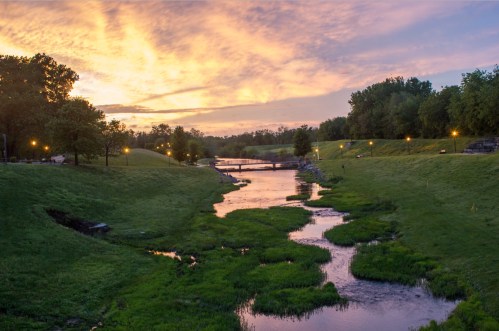 A serene landscape at sunset, featuring a winding stream, lush green banks, and a colorful sky.