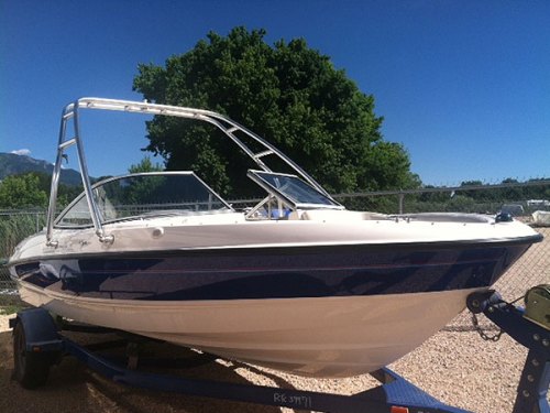 A white and blue boat on a trailer, set against a clear blue sky and green trees in the background.
