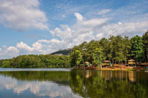 A serene lake surrounded by lush trees and a distant mountain under a partly cloudy sky. Reflections on the water.