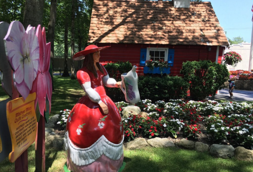 A colorful garden scene featuring a statue of a woman in a red dress near a quaint red house and vibrant flowers.