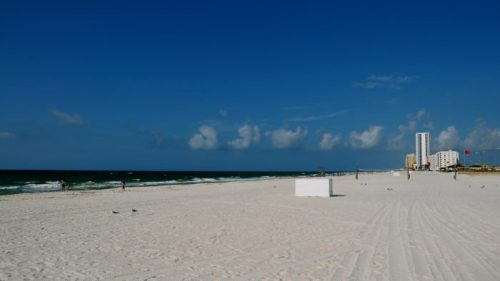A sunny beach scene with white sand, gentle waves, and a clear blue sky, featuring distant buildings along the shore.