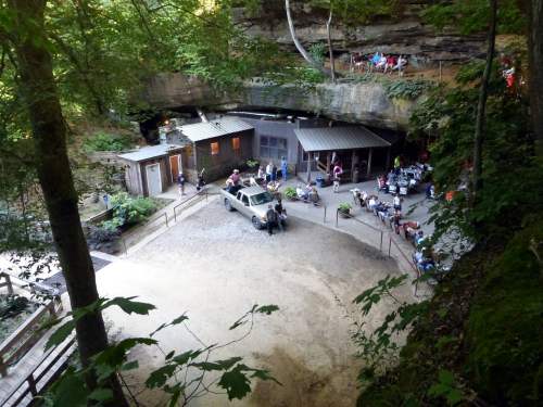A scenic view of a cave entrance with a restaurant patio, surrounded by trees and visitors enjoying the outdoors.
