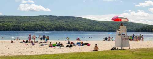 A sandy beach with people sunbathing and swimming, a lifeguard tower in the foreground, and green hills in the background.