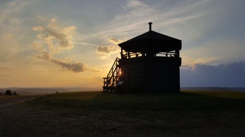 Silhouette of a lookout tower against a sunset sky, with clouds and rolling hills in the background.