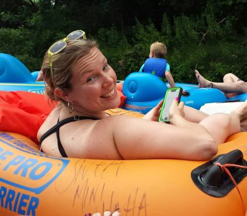 A woman smiles while relaxing on an inflatable raft, holding a phone, with others in the background on a river.