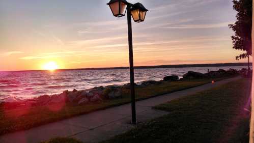 A sunset over a lake, with a lamp post and rocky shoreline along a grassy path.