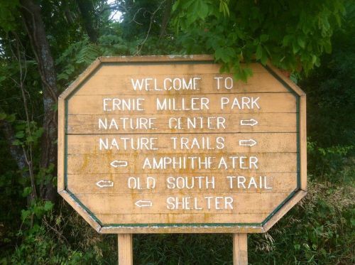 Wooden sign welcoming visitors to Ernie Miller Park, listing directions to the Nature Center, trails, amphitheater, and shelter.