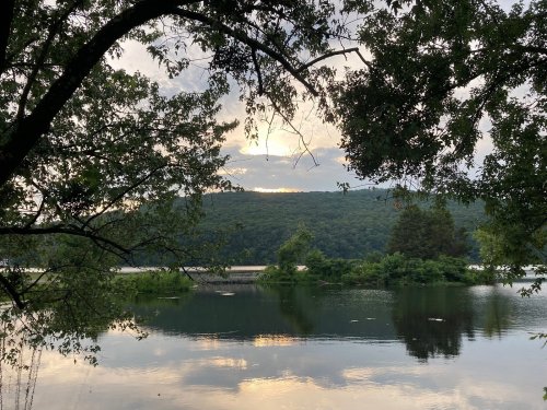 A serene lake scene framed by trees, with a cloudy sky and distant hills reflecting on the water's surface.