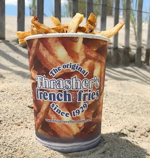 A cup of Thrasher's French fries sits on sandy beach, with wooden fence and blue sky in the background.