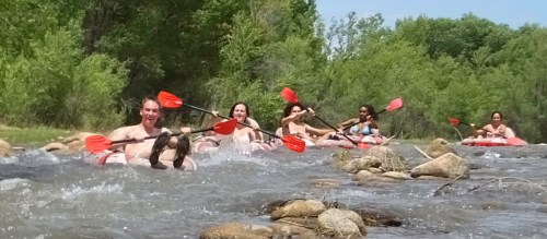 A group of people kayaking down a river, surrounded by greenery and rocks, enjoying a sunny day outdoors.