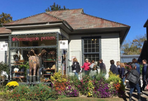 A line of people waiting outside a pottery shop with colorful flowers in front and a clear blue sky above.