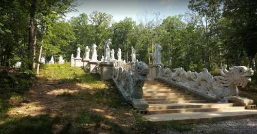 A stone pathway leads up to intricately carved statues in a wooded area, surrounded by greenery.