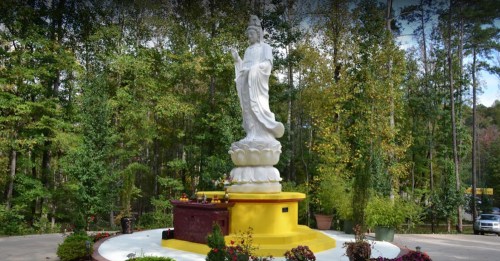 A tall white statue of a figure stands on a yellow pedestal, surrounded by greenery and colorful flowers.