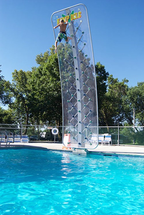 A person climbs a tall, curved rock wall above a swimming pool on a sunny day, surrounded by trees.