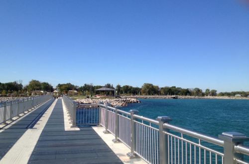 A scenic view of a pier extending over blue water, surrounded by trees and a gazebo in the distance under a clear sky.