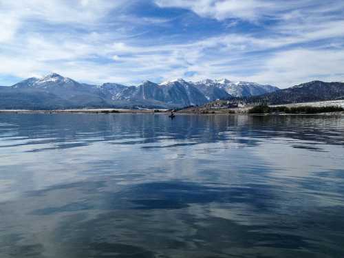 A serene lake reflects snow-capped mountains and a blue sky with wispy clouds.