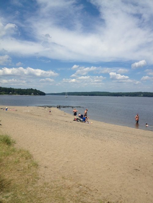 A sandy beach with people relaxing and swimming, under a blue sky with fluffy clouds and a calm river in the background.