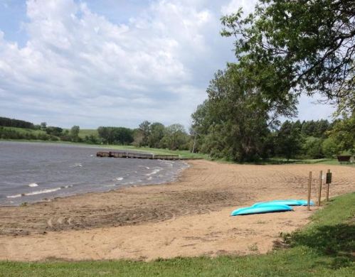 A sandy beach by a calm lake, with a dock in the distance and trees lining the shore under a cloudy sky.