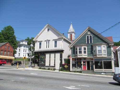Two historic buildings in a small town, one with a green facade and the other white, under a clear blue sky.
