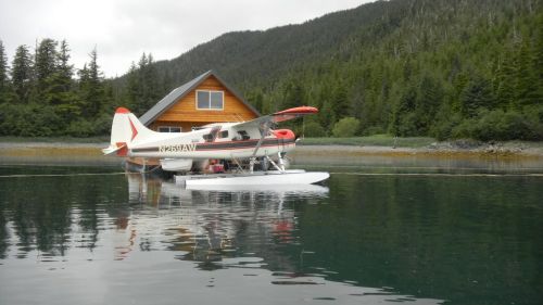 A seaplane docked beside a wooden cabin on a calm lake, surrounded by lush green trees and mountains.