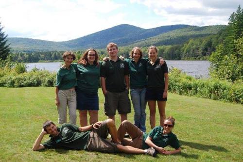 A group of seven people in matching green shirts poses outdoors by a lake, with mountains in the background.