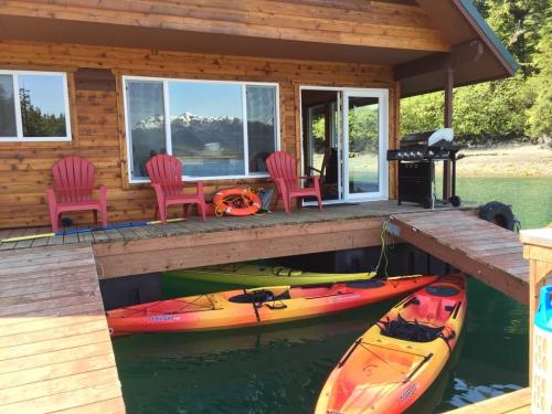 A wooden dock with two kayaks, a grill, and chairs by a waterfront cabin surrounded by greenery and mountains.